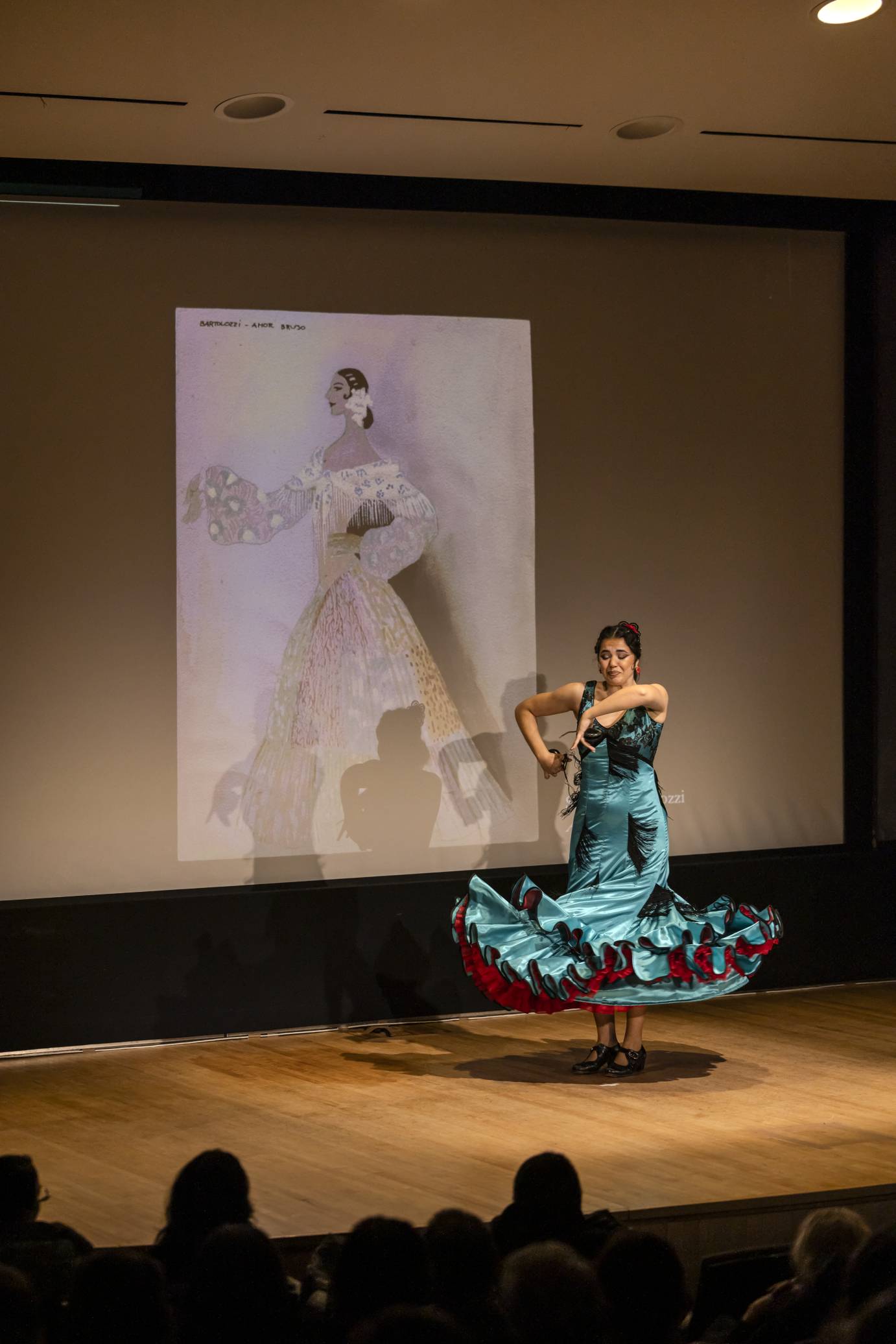 A flamenco dancer swirls her shiny aqua ruffled skirt tightly around her legs. Her elbows are lifted as she plays the castanets looking down.